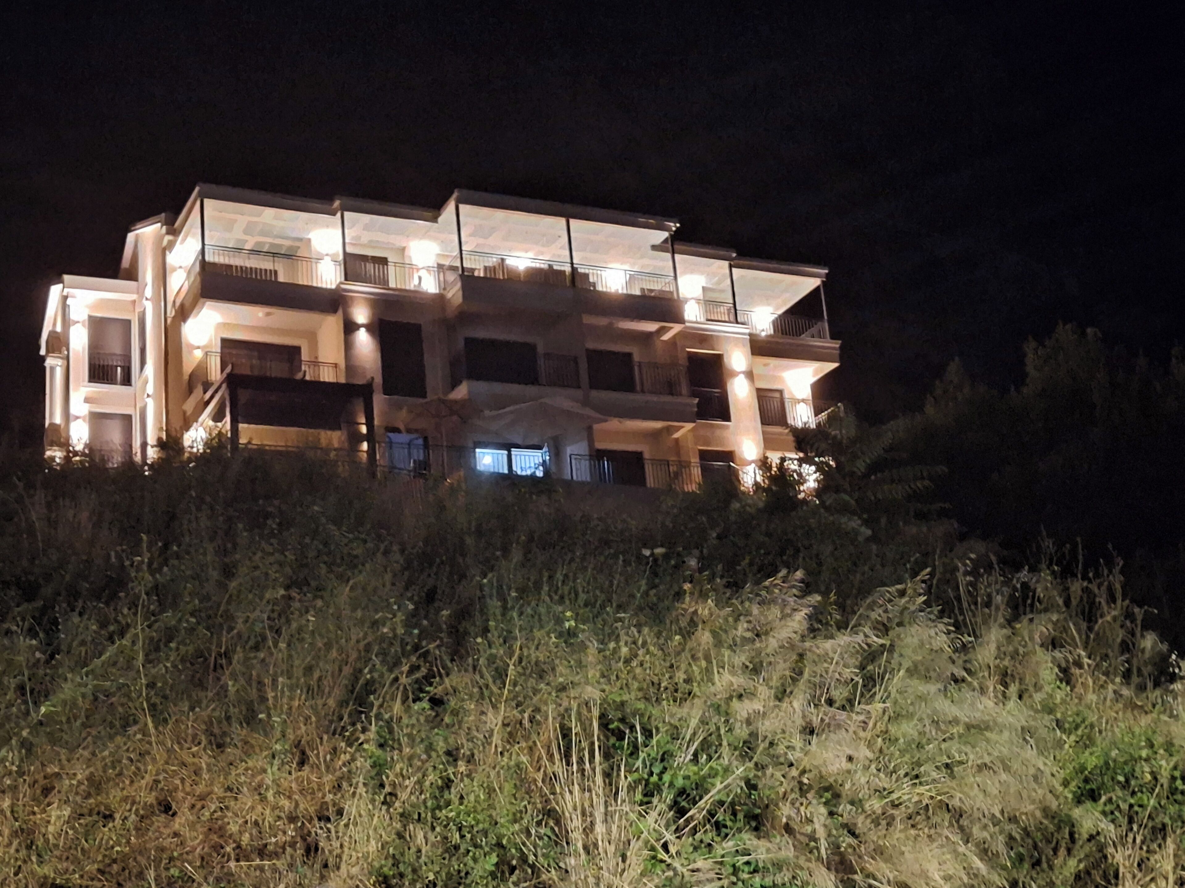 A multi-story building with balconies and a flat roof, surrounded by vegetation.