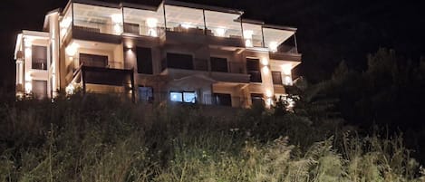 A multi-story building with balconies and a flat roof, surrounded by vegetation.