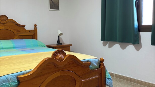 A bedroom with a wooden bed, a bedside table, a nightstand, a wall-mounted picture, and a window with curtains. - STAR OF THE SOUTH (Valle La Lajita)
