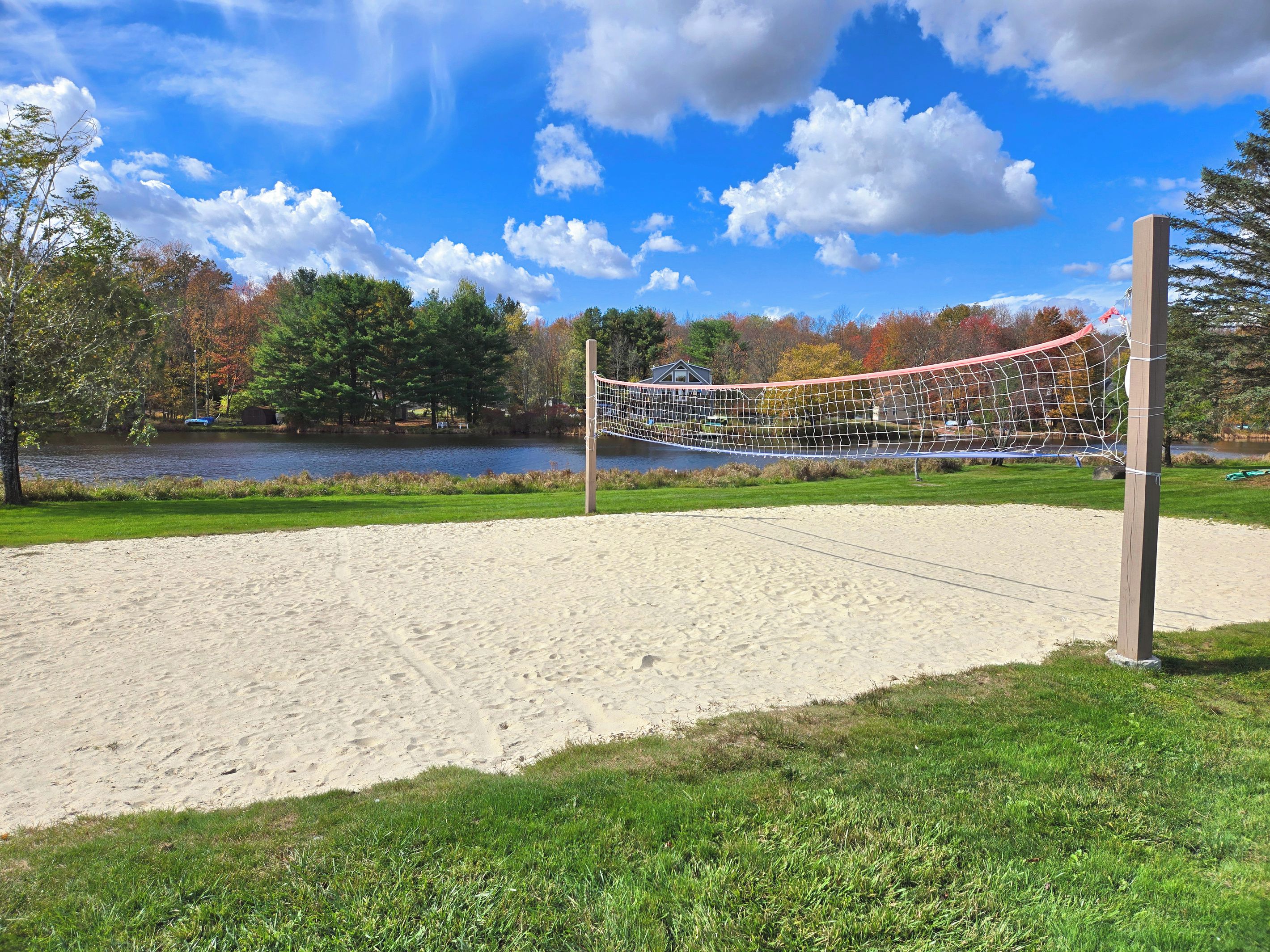 Una cancha de voleibol de arena con red, rodeada de césped y árboles, con un lago y una casa al fondo.