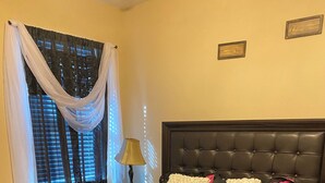 A bedroom with a dark-colored bed, a nightstand with a lamp, and a window with sheer curtains. - CLOSE TO DISNEY Cozy 3 bed/3 bed house in tranquil Apopka w fitness room/ pool (Apopka)