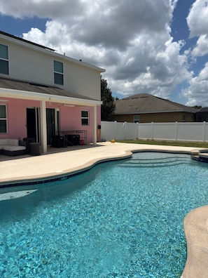 Ein Pool mit geschwungener Kante, eine Terrasse mit weißem Zaun und ein zweistöckiges Haus mit rosa Fassade.