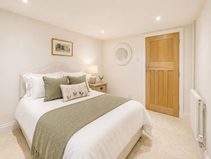A bedroom with a bed, pillows, a bedside table with a lamp, a round mirror, and a wooden door. - The Hyde | Private Hot tub & Pool | Modern (Prestbury)