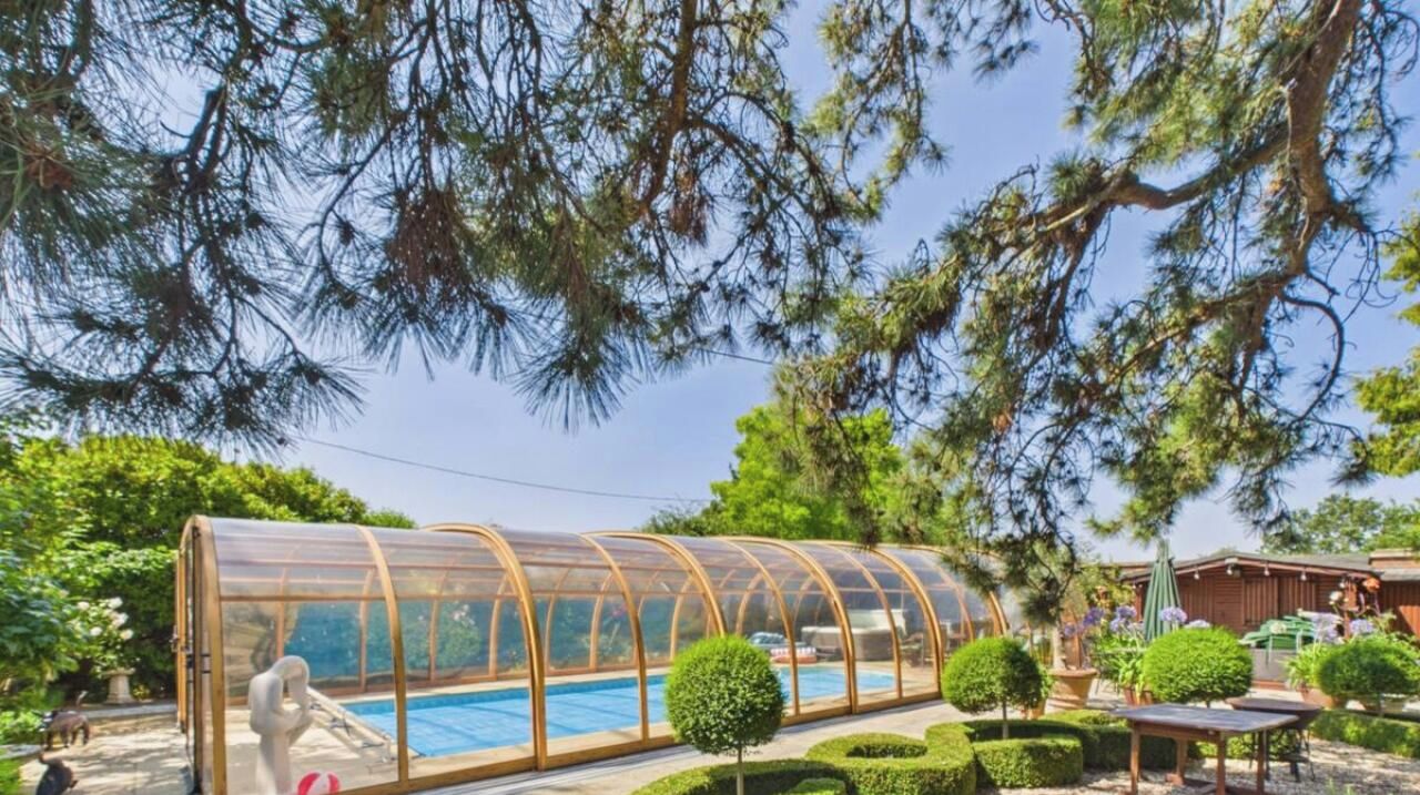 A covered swimming pool with a transparent roof, surrounded by neatly trimmed bushes and trees.