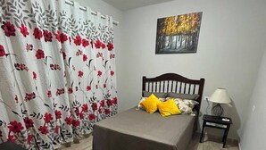 A bedroom with a bed, a nightstand, a lamp, a painting on the wall, and a window with curtains. - Vrbo Property (Araçariguama)