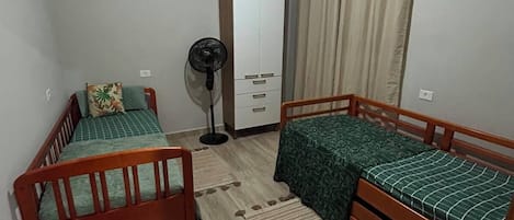 Una habitación pequeña y luminosa con dos camas de madera, un armario, un ventilador y una ventana con cortinas.