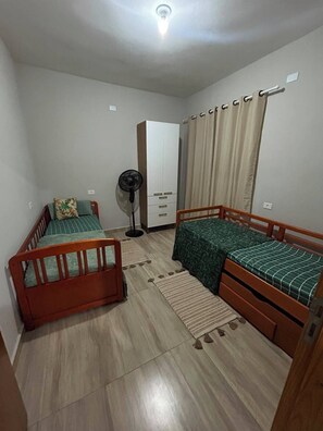 Une petite pièce bien éclairée, avec deux lits en bois, une armoire, un ventilateur et une fenêtre avec des rideaux.