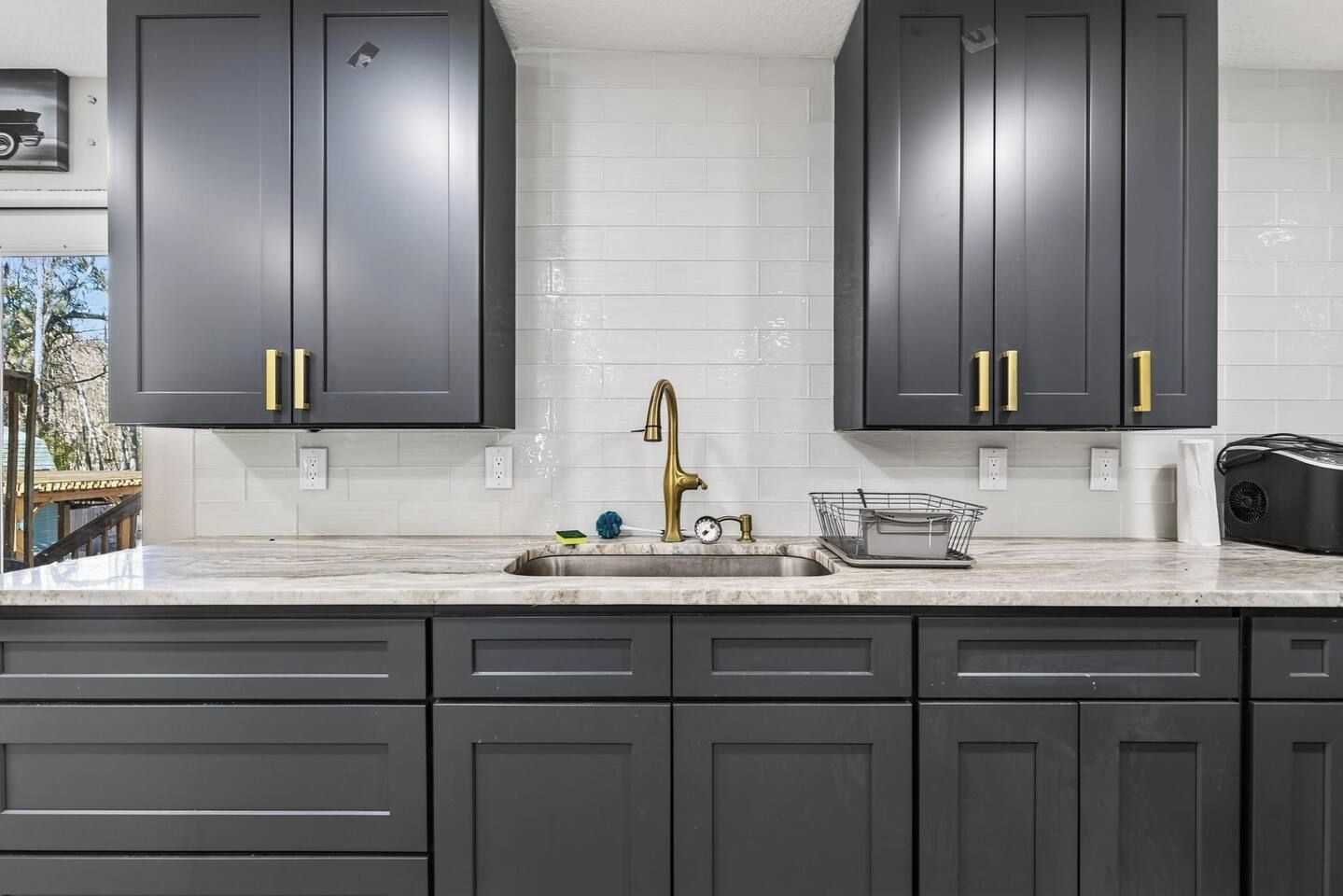 A kitchen with dark cabinets, a white tiled backsplash, a stainless steel sink, and a gold faucet.
