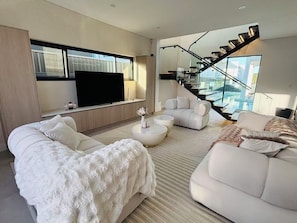 A modern living room with a flat-screen TV, two sofas, a coffee table, and a staircase leading to a balcony with a pool view. - Spacious 5BR Home, Private Pool Near Syd Airport (Bexley)