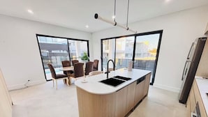 A modern kitchen with a central island, a dining area with chairs, and large windows. - Spacious 5BR Home, Private Pool Near Syd Airport (Bexley)