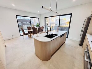 A modern kitchen with a central island, a dining area with chairs, and large windows. - Spacious 5BR Home, Private Pool Near Syd Airport (Bexley)
