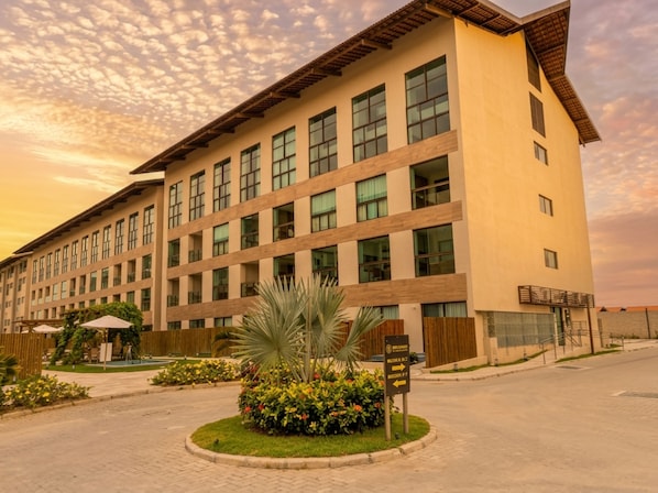 Ein mehrstöckiges Hotel mit flachem Dach, großen Fenstern und einem begrünten Eingangsbereich mit einer Palme und Blumenbeeten.