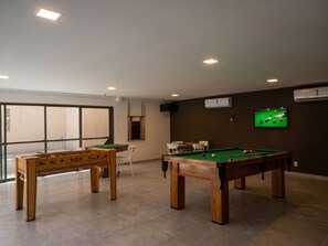 Ein Raum mit Billardtisch, Kicker und Poolbillard. An der Wand befinden sich Klimaanlagen und ein Fernseher zeigt ein Billardspiel.