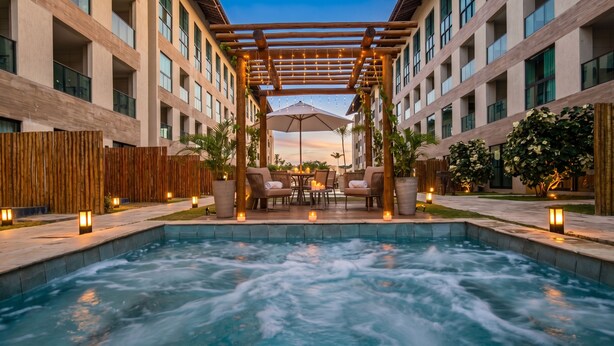 Zona de piscina de hotel con hidromasaje, sillones de descanso y una pérgola con luces colgantes.