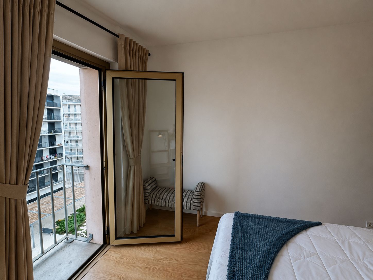 A bedroom with a bed, a window with curtains, a view of a balcony, and a door leading to another room.
