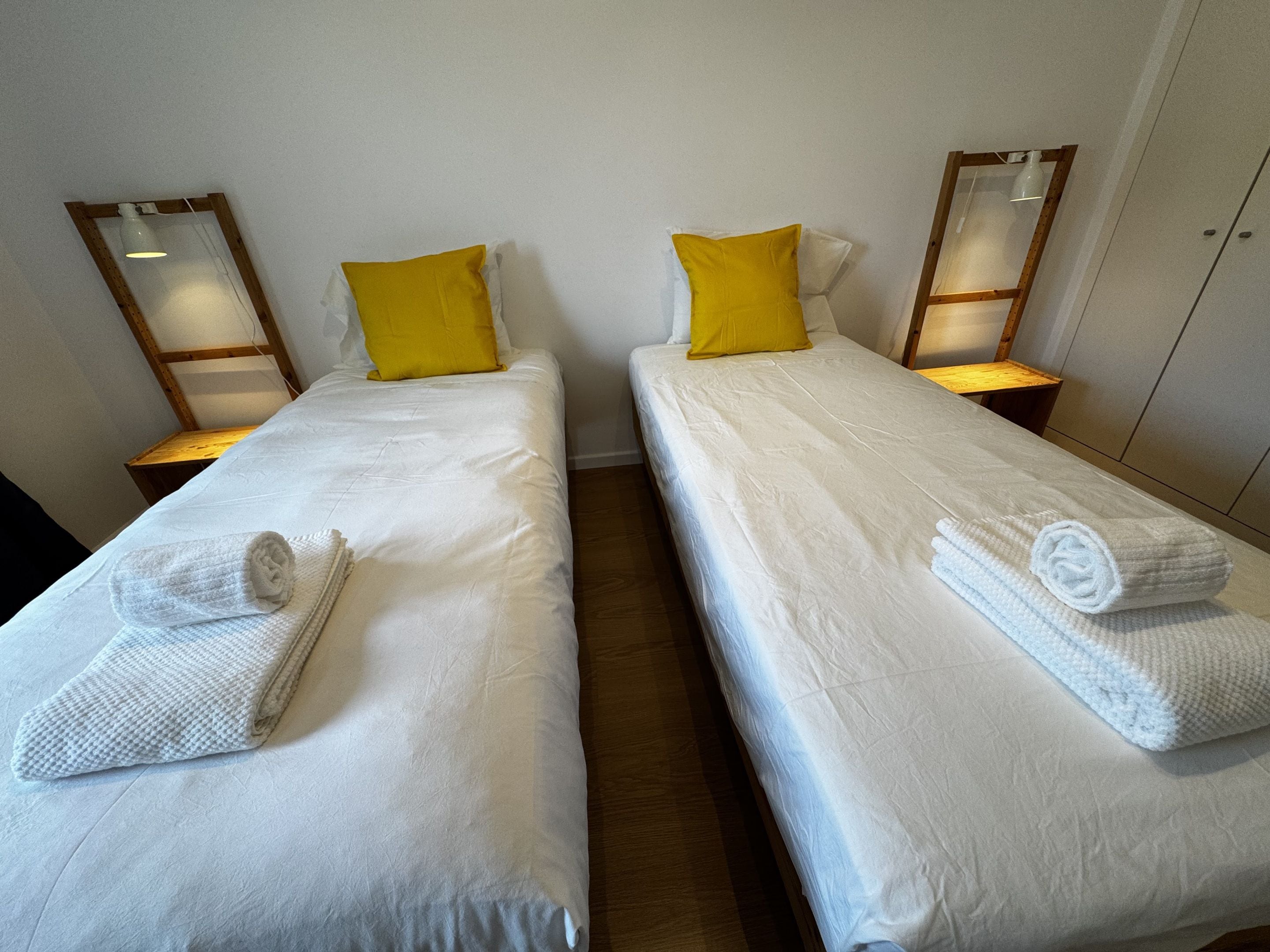 Deux lits simples avec du linge de lit blanc et des oreillers jaunes, chacun agrémenté d’une serviette roulée et d’une table de chevet avec une lampe.