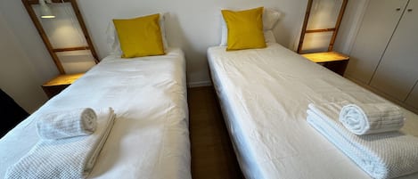 Deux lits simples avec du linge de lit blanc et des oreillers jaunes, chacun agrémenté d’une serviette roulée et d’une table de chevet avec une lampe.