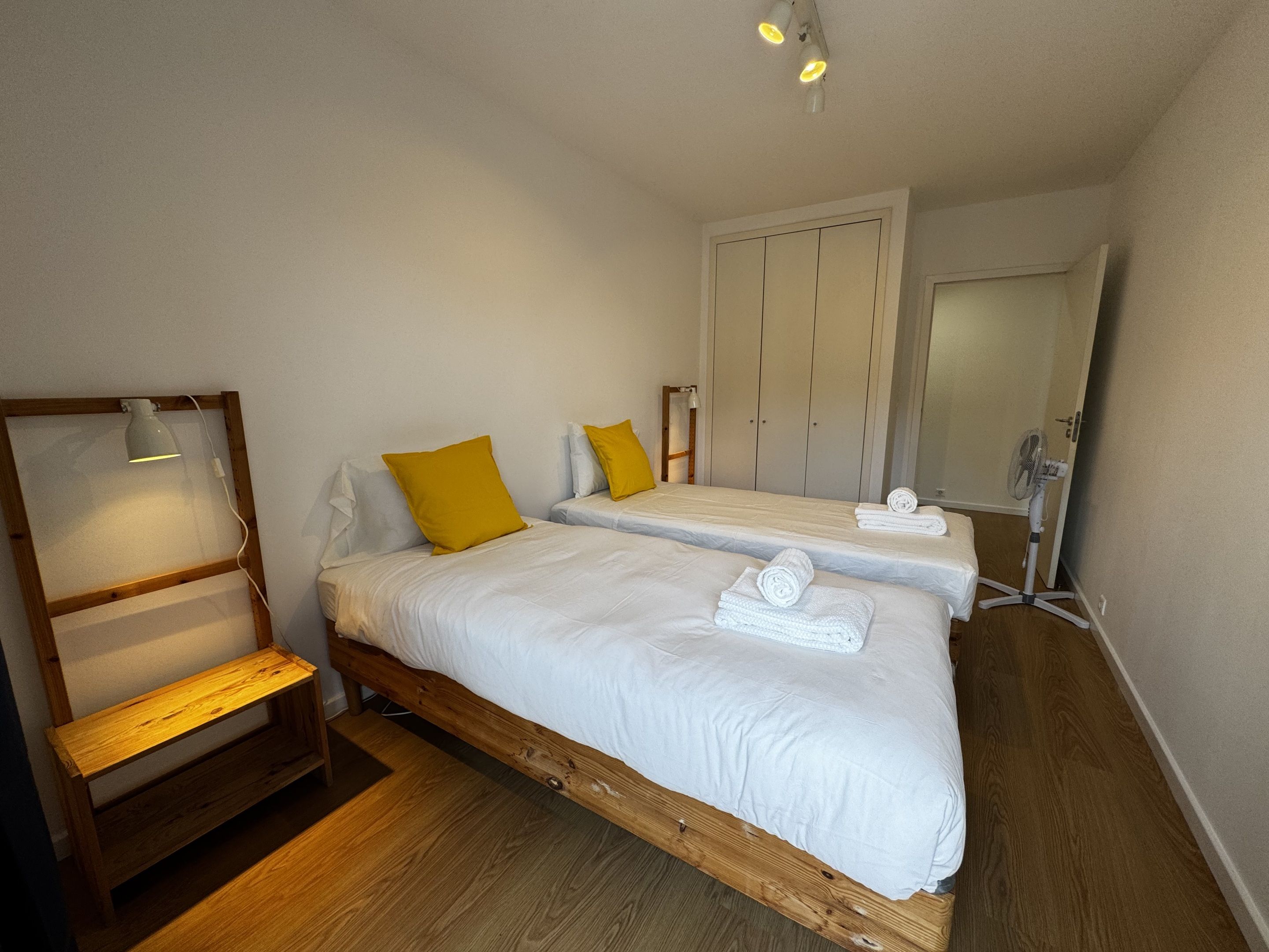 A bedroom with two beds, a wooden bench, a desk with a lamp, and a wardrobe.