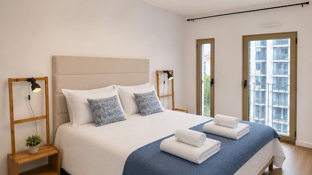 A modern bedroom with a large bed, a bedside table with a lamp, and a view of the outside through a window. - Lisbon Green Center (Lisboa)