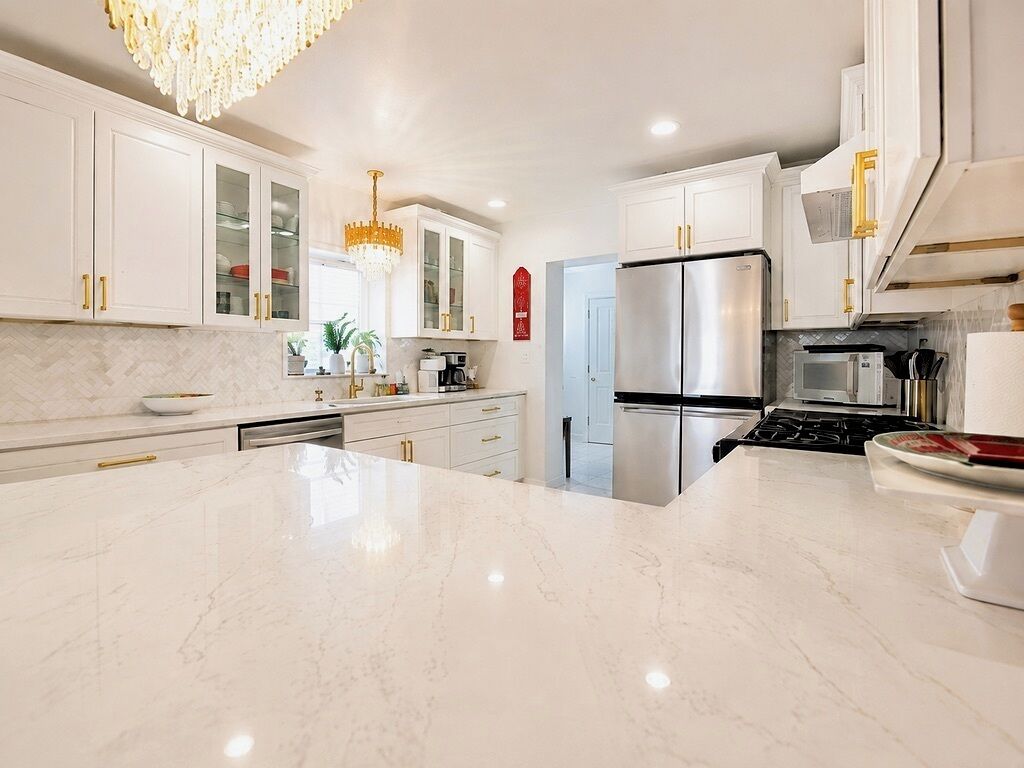 A modern kitchen with white cabinetry, a large marble countertop, stainless steel appliances, and a chandelier.