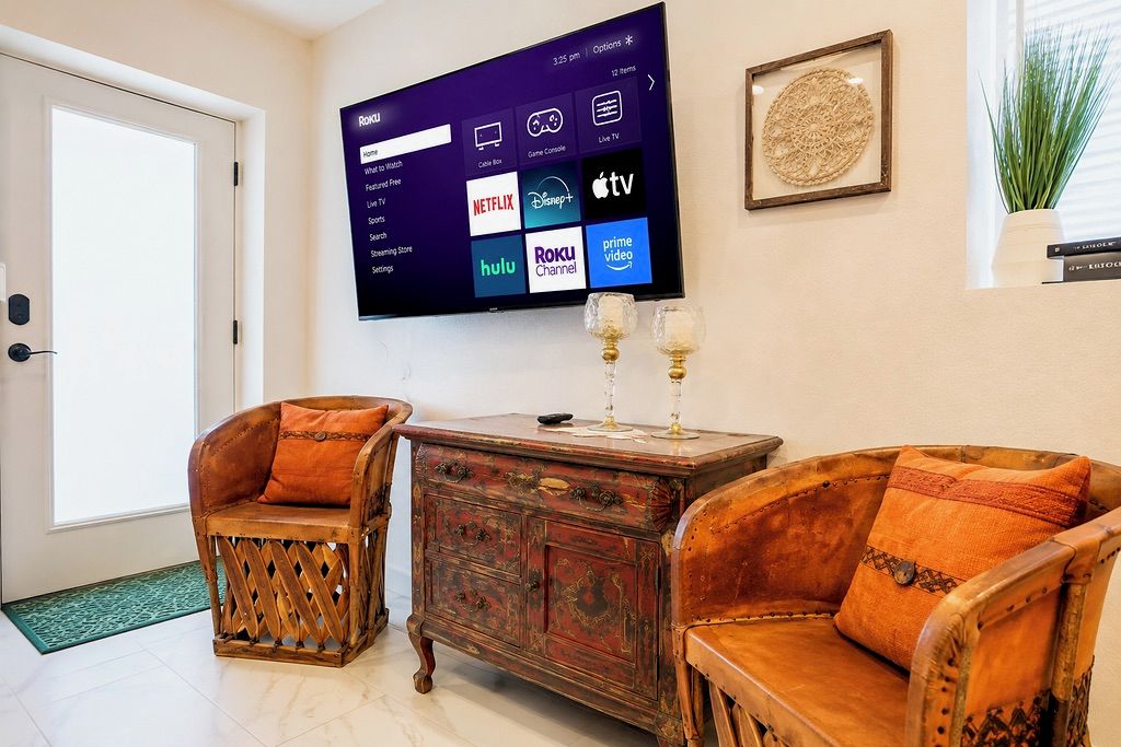 Ein Wohnzimmer mit einem Flachbildfernseher, der Streaming-Dienste anzeigt, einer Holzvitrine mit Gläsern und zwei Polstersesseln.