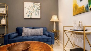 Un divano blu con un cuscino decorato, un tavolino in legno, un comodino dorato con una lampada e un quadro appeso al muro.