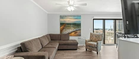 A living room with a sectional sofa, a patterned armchair, a glass coffee table, and a painting of a beach scene.