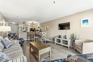 A living room with a sofa, coffee table, TV, and a dining area with chairs and a table. - Shorewalk 5- 2 Bedroom- Beach Across The Street (North Myrtle Beach)