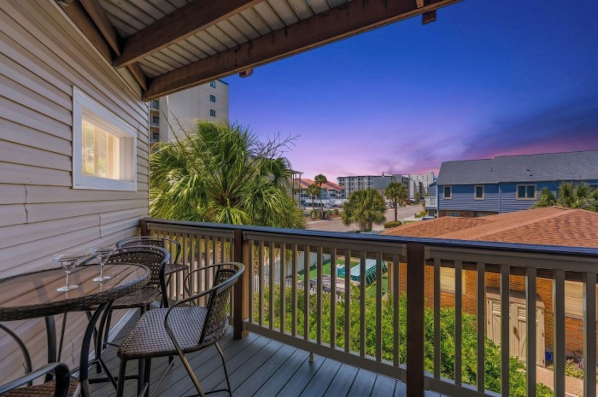 A balcony with a table and chairs, overlooking a residential area with houses and a clear sky.