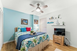 A bedroom with a bed, a ceiling fan, a TV, and a shelf with decorative items. - Shorewalk 5- 2 Bedroom- Beach Across The Street (North Myrtle Beach)