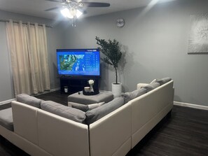 Una sala de estar con un sofá modular, un televisor de pantalla plana, una planta en maceta, un reloj de pared y un ventilador de techo.