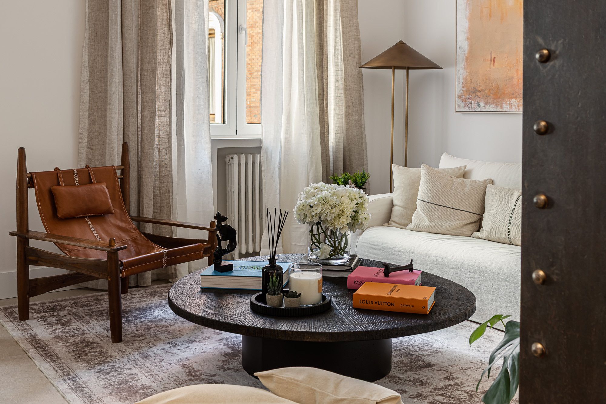 A living room with a leather chair, a round coffee table with books and a candle, a white sofa, and a floor lamp.