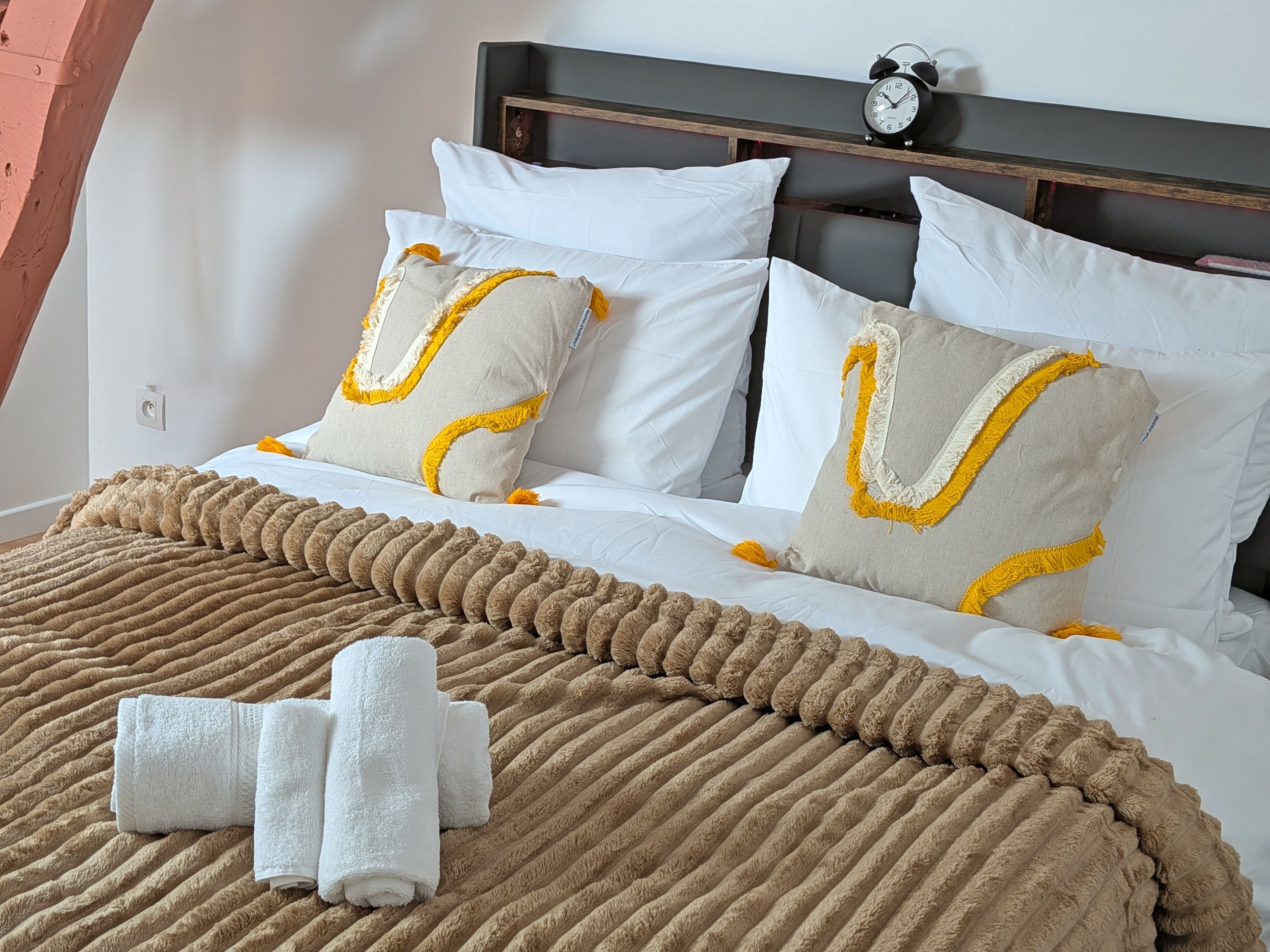 A neatly made bed with white and beige pillows, a brown textured blanket, and two rolled white towels.