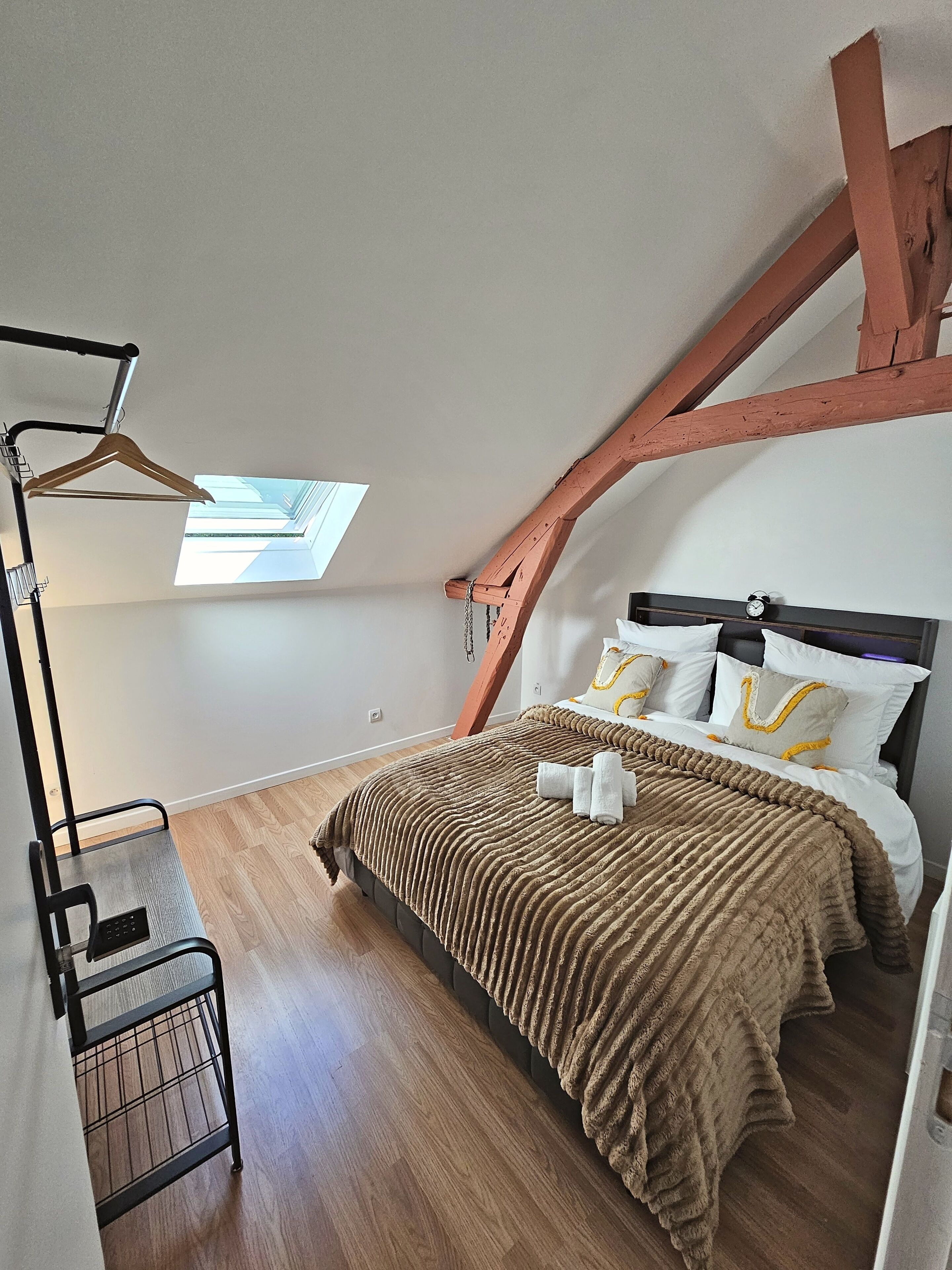 A bedroom with a sloped ceiling, a bed with a brown blanket, a hanging light fixture, and a small table with a chair.