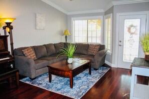 A living room with a sectional sofa, a coffee table, a piano, and a door with a decorative wreath.