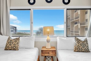 Ein Hotelzimmer mit zwei Betten, einem Nachttisch mit Lampe und Blick auf Strand und Gebäude.