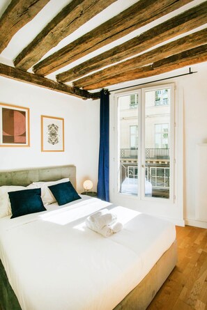 Ein Schlafzimmer mit Bett, Kopfstütze, Nachttisch, Fenster mit Blick auf einen Balkon und Holzbalken an der Decke.