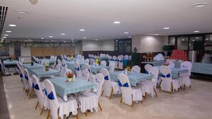 Uma sala de banquetes com várias mesas e cadeiras, cada uma com toalha azul e cadeiras brancas adornadas com fitas azuis.