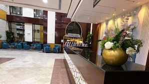Uma área de recepção de hotel com piso de mármore, assentos azuis, balcão de recepção e parede decorativa com plantas.