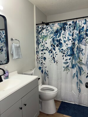 Un baño con un lavamanos blanco, un inodoro, una cortina de ducha con estampado floral y una alfombra de baño azul.