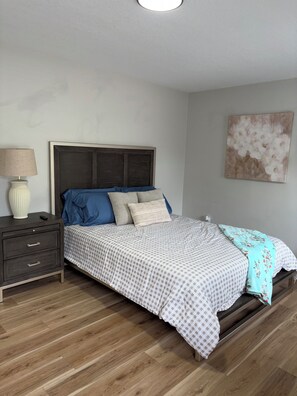 Un dormitorio con cama, mesita de noche, lámpara y una obra de arte en la pared.