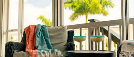 A wicker chair with a striped throw blanket, a small round table with two drinks, and a view of palm trees through large windows.
