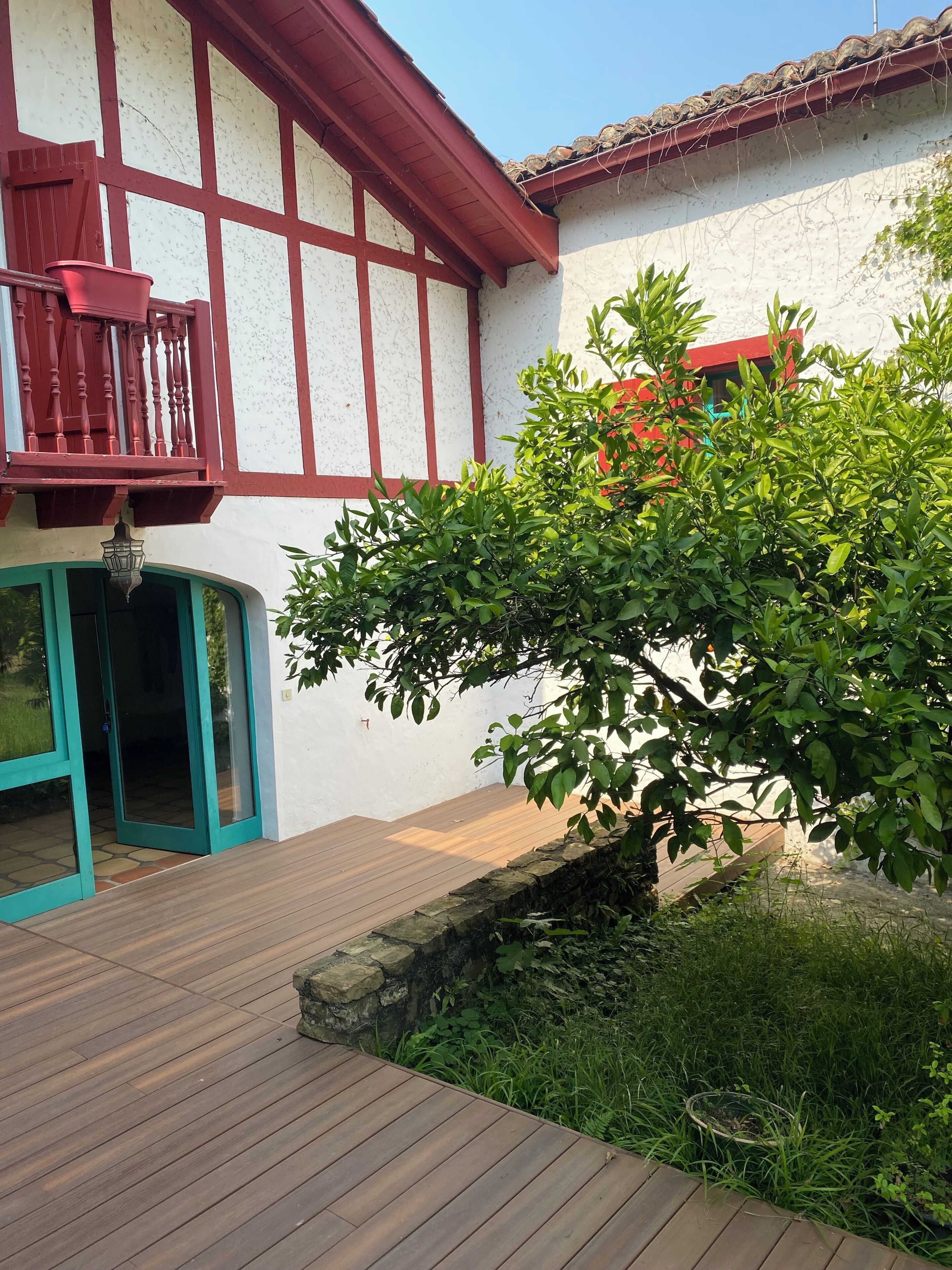 Une terrasse en bois mène à un bâtiment blanc agrémenté de touches de rouge, avec une chaise rouge et une porte verte.
