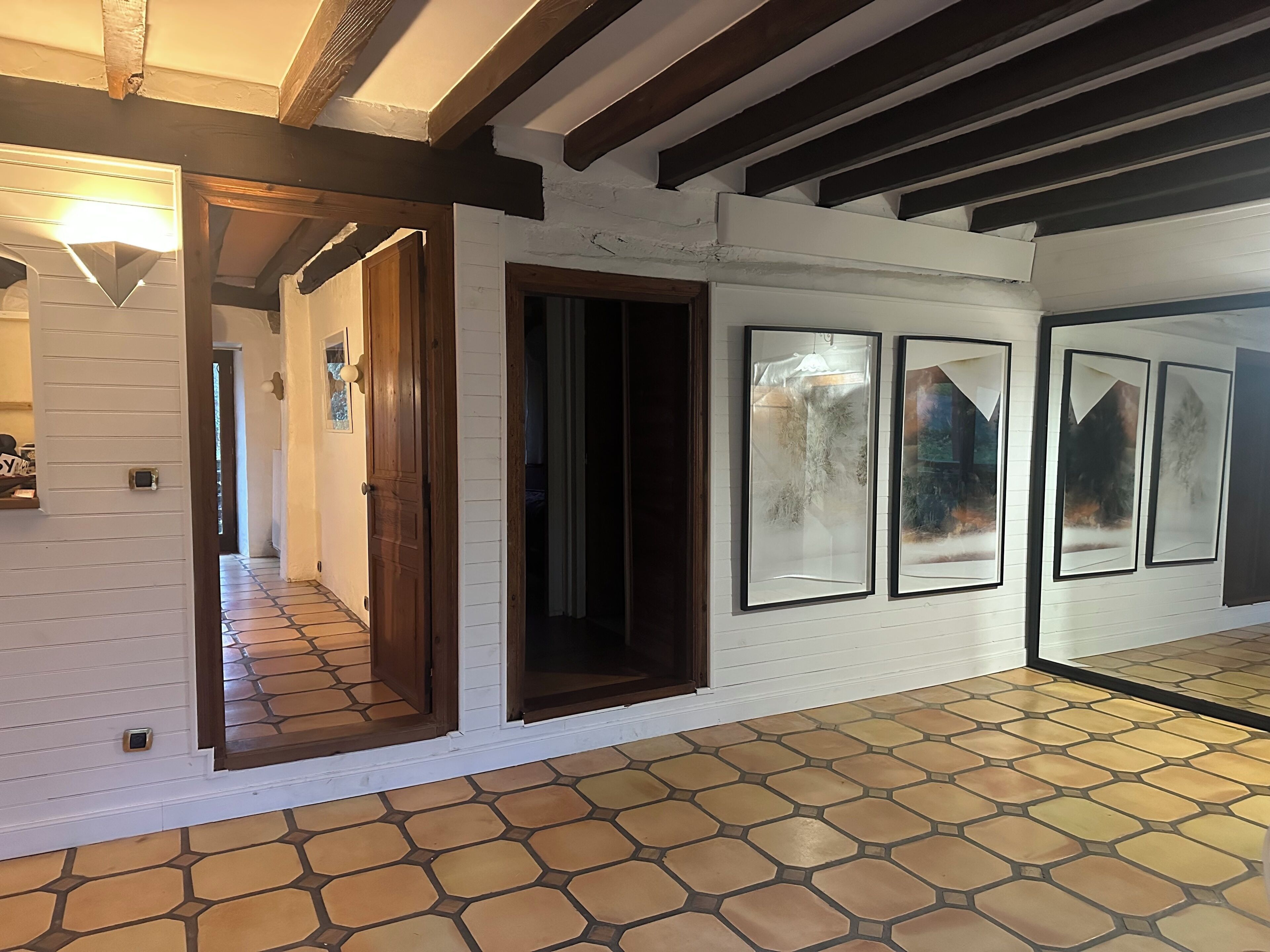 Un couloir aux murs blancs, doté d’un sol carrelé et de poutres en bois au plafond. On y trouve des portes menant à d’autres pièces, ainsi qu’un mur orné de tableaux encadrés.