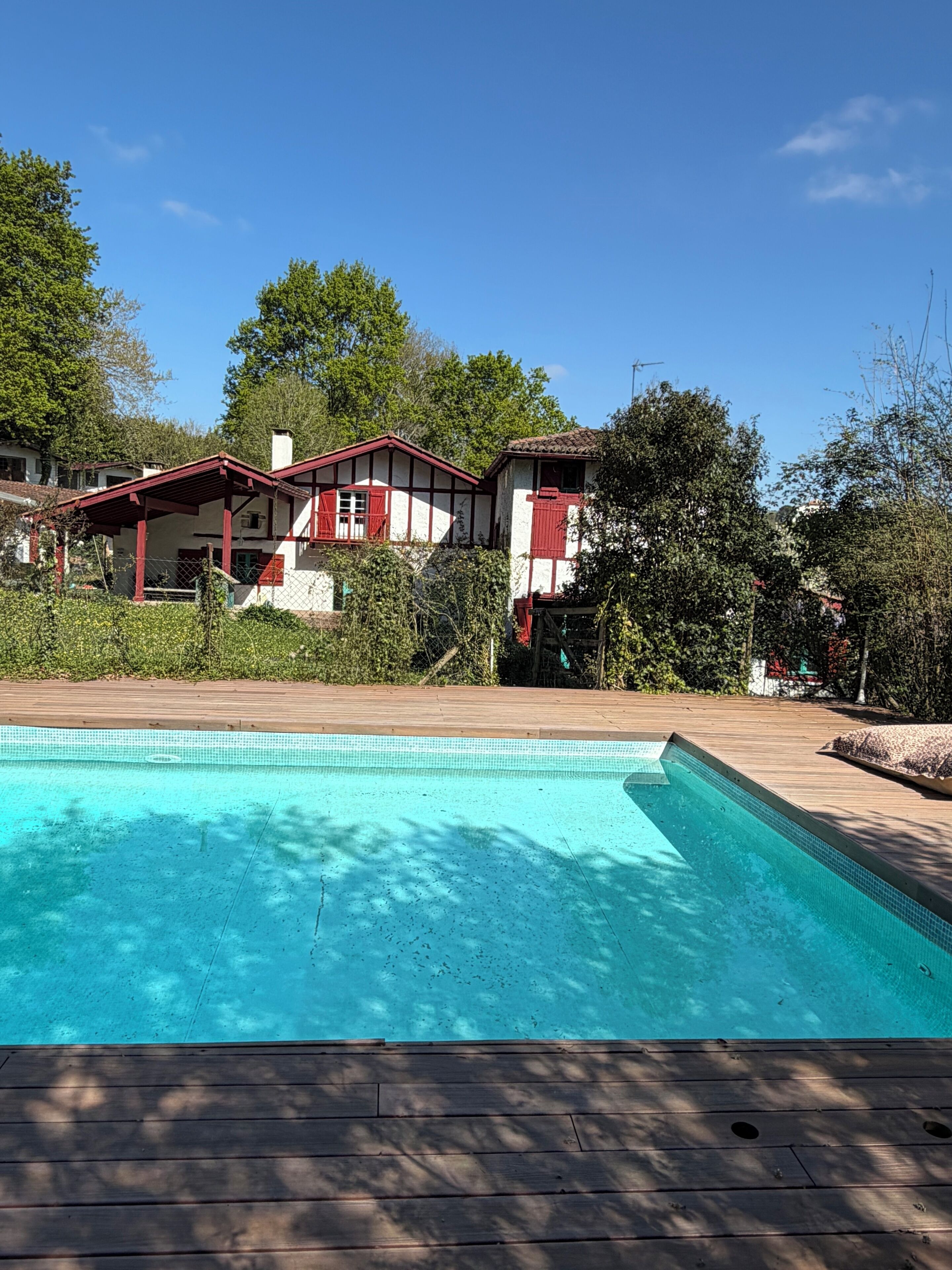 Une piscine avec une terrasse en bois, entourée d’une maison à la façade rouge et blanche, et des arbres en arrière-plan.