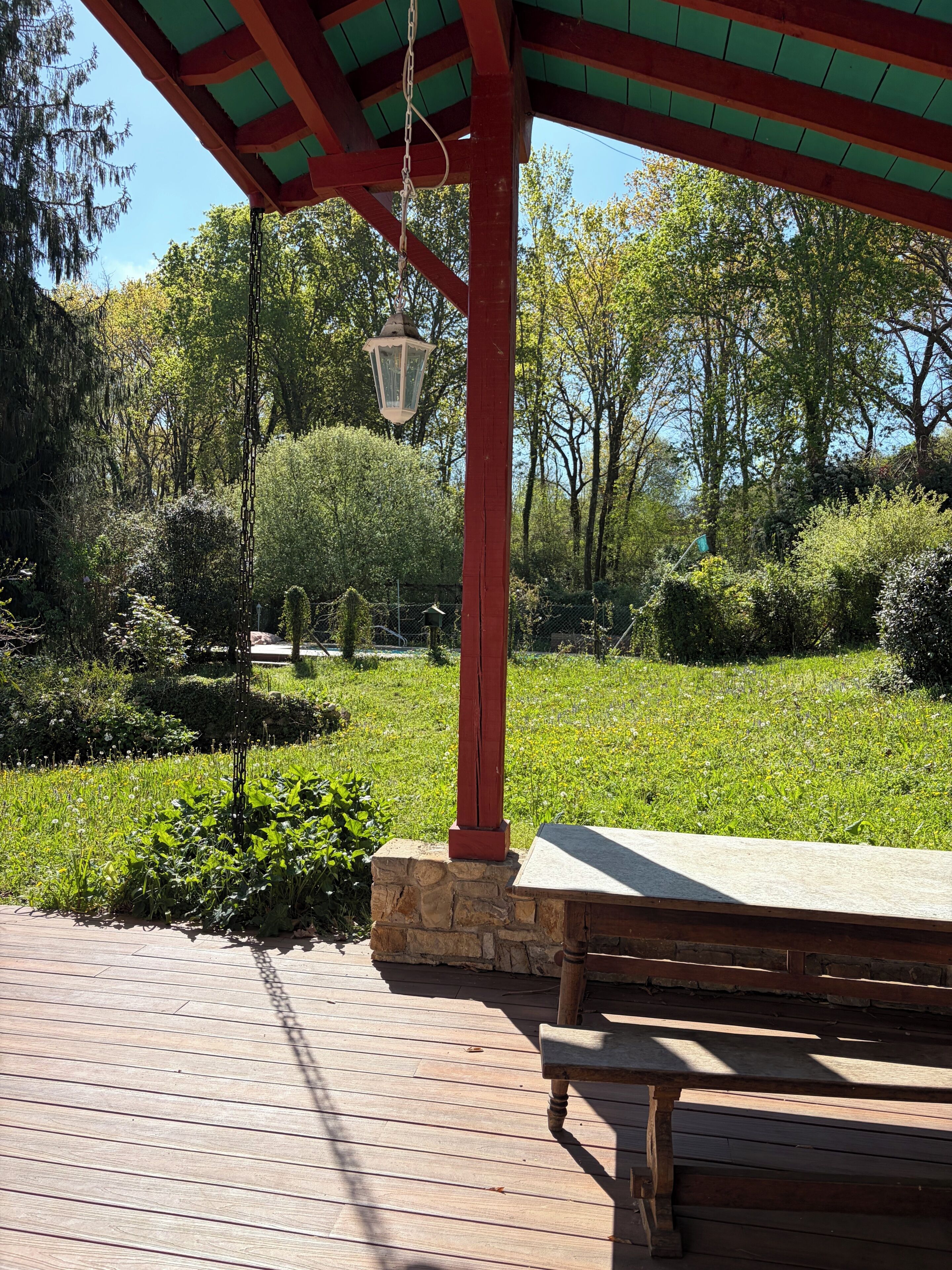 Een houten bank onder een dak dat rood en groen is, omringd door een tuin met diverse planten en bomen.