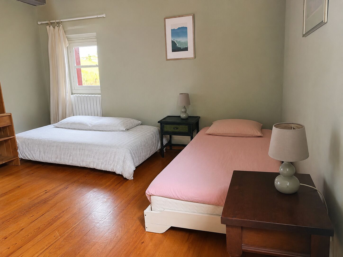 Een slaapkamer met twee bedden, een raam met gordijnen, een nachtkastje met een lamp en een lijst met een afbeelding aan de muur.