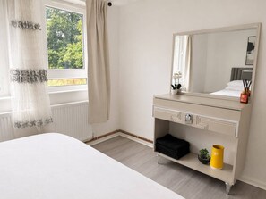 A bedroom with a bed, a bedside table with a mirror, a window with curtains, and a radiator. - Near Wembley Stadium- 1 Bedroom Flat (Wembley)