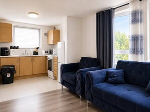 A modern kitchen with wooden cabinets, a black trash bin, and a blue sofa set. - Near Wembley Stadium- 1 Bedroom Flat (Wembley)