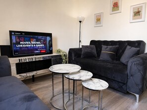 Una sala de estar moderna con un televisor de pantalla plana mostrando un servicio de streaming, un sofá negro y dos mesas auxiliares con cubierta de mármol.
